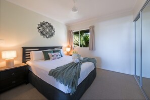 A bedroom with a bed, bedside tables, a window with curtains, and a decorative wall piece. - Waterlily - Two Bedroom Apartment (Airlie Beach)