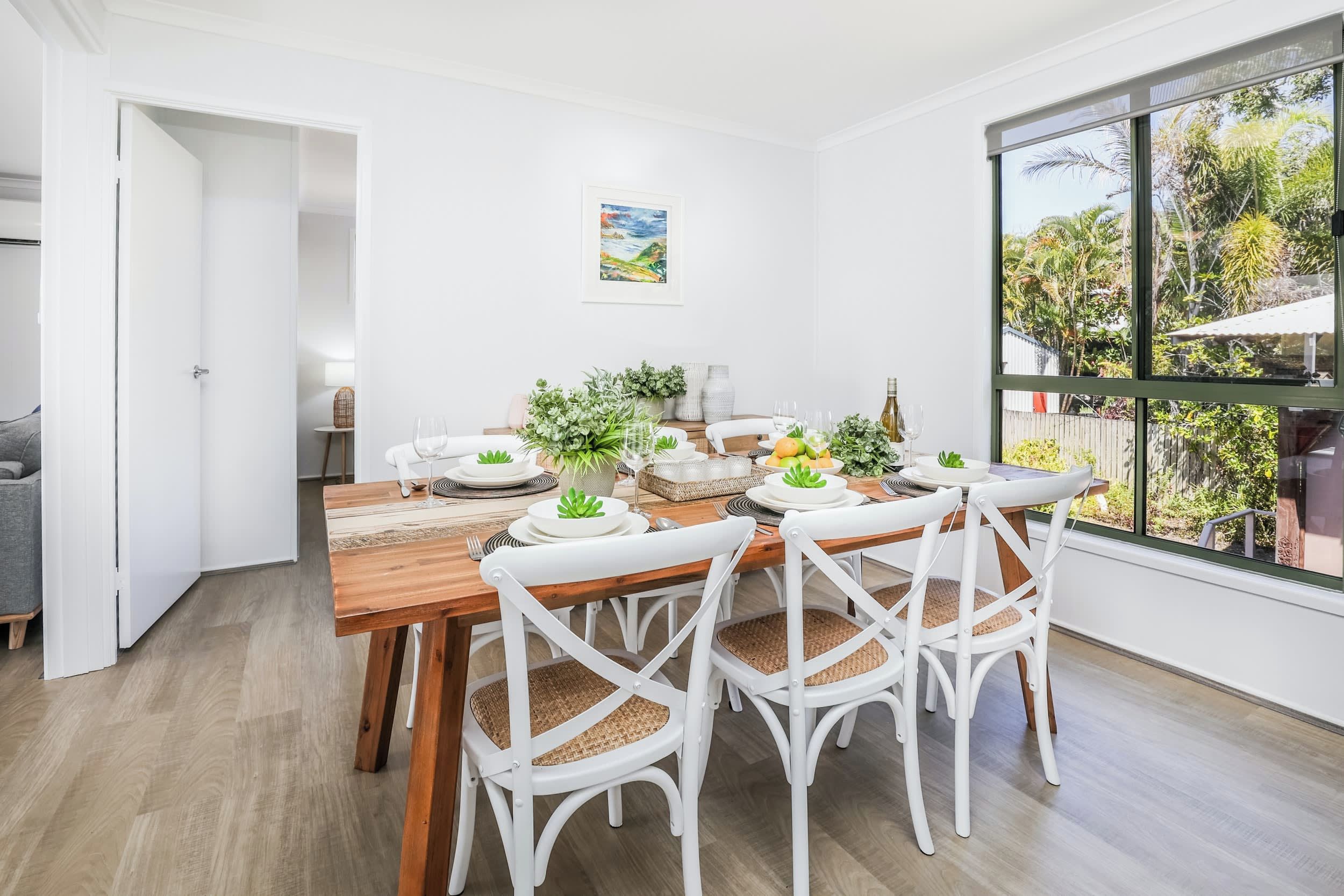 A dining area with a wooden table set for four, white chairs, and a view of greenery outside.