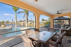 Un patio soleado con una mesa de vidrio y sillas de mimbre, con vistas a una piscina y una zona residencial.