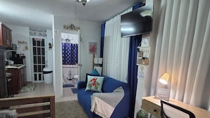 Un espace de vie compact comprenant un coin cuisine, un canapé agrémenté de coussins décoratifs, une télévision, un bureau avec une chaise et une salle de bain avec un rideau de douche.