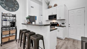 A modern kitchen with white cabinetry, a central island with stools, a large clock, and a microwave. - The Silo Sweetheart- Just minutes from Downtown! (Waco)