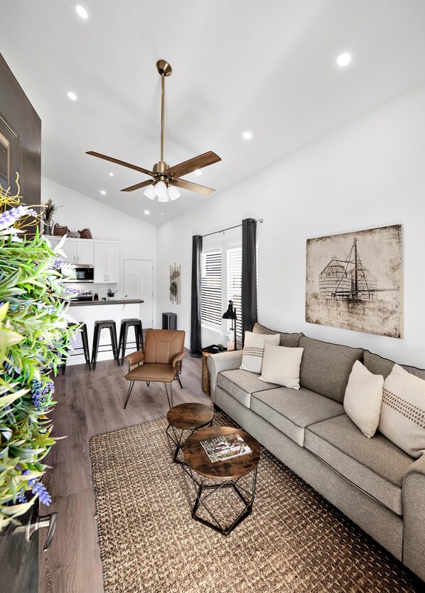 A modern living room with a sofa, coffee table, and chairs, a kitchen area with white cabinets, and a ceiling fan.