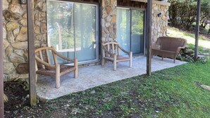 En fasad av sten med en veranda av trä, två fåtöljer av rotting och en bänk.