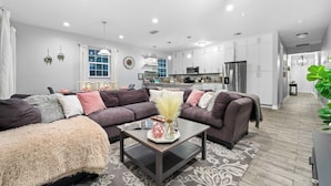 A modern living room with a sectional sofa, a coffee table, and a kitchen in the background. - Pet-Friendly Home with a Hot Tub, Climbing Wall, King Bed & Gym by Mane Suites! (Tampa)