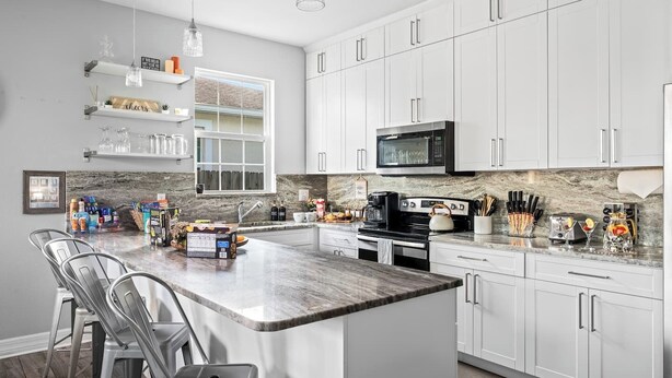 A modern kitchen with white cabinets, a granite countertop, stainless steel appliances, and bar stools. - Pet-Friendly Home with a Hot Tub, Climbing Wall, King Bed & Gym by Mane Suites! (Tampa)