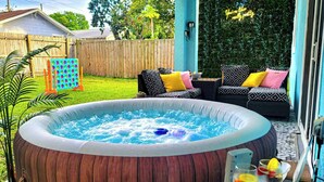 An outdoor hot tub area with a wooden deck, seating with cushions, and a glass table with drinks. - Pet-Friendly Home with a Hot Tub, Climbing Wall, King Bed & Gym by Mane Suites! (Tampa)
