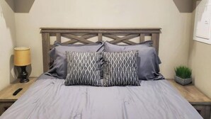 Ein Bett mit Holz-Kopfteil, zwei Kissen mit geometrischem Muster, eine Nachttischlampe und ein kleiner Topfpflanze.