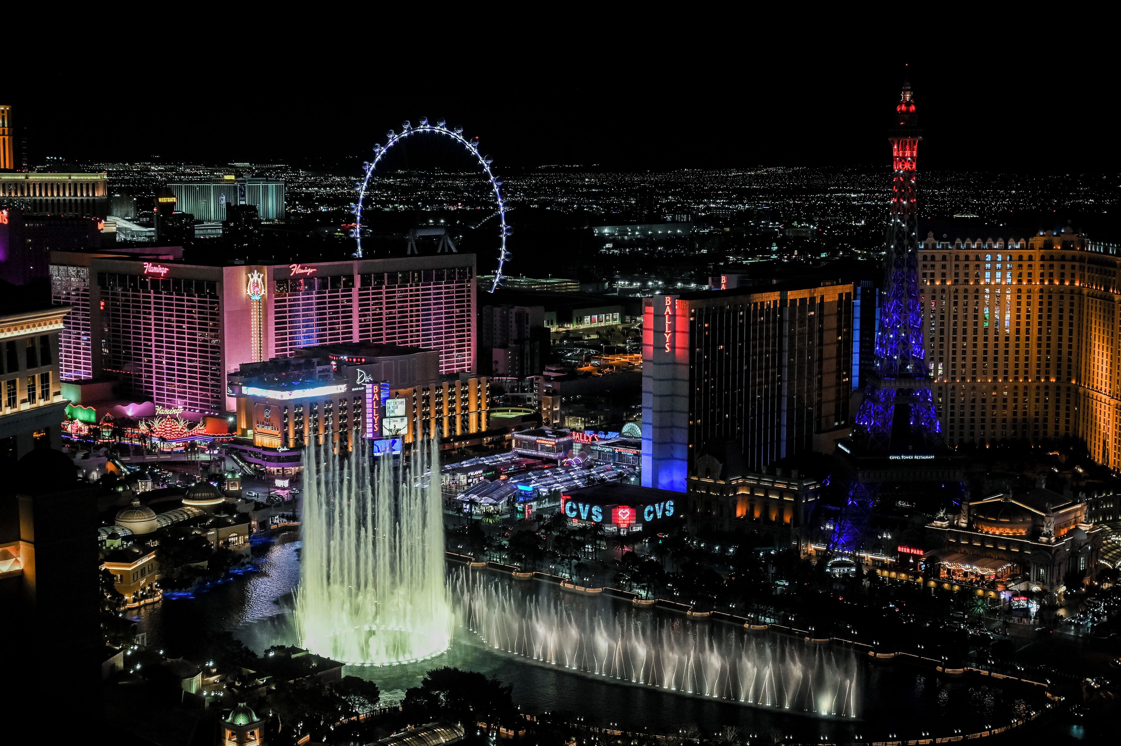 A nighttime cityscape with illuminated buildings, a prominent Ferris wheel, and a large fountain.