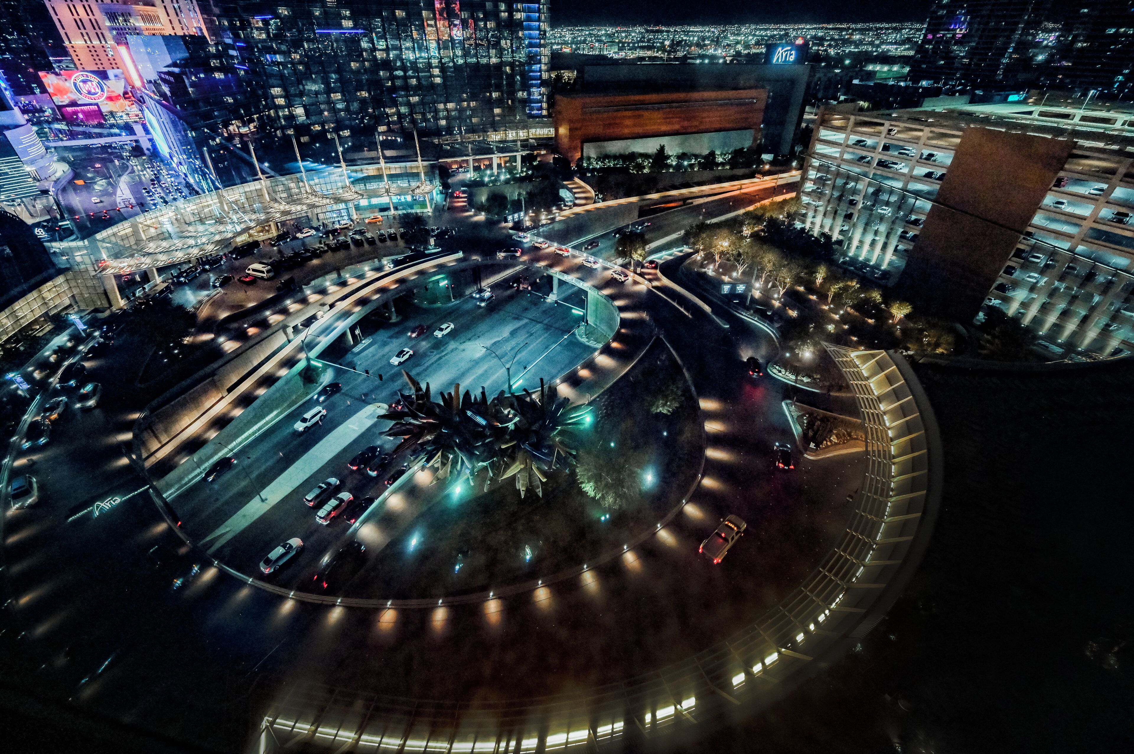An aerial view of a roundabout with multiple lanes of traffic, surrounded by illuminated buildings and a parking structure.