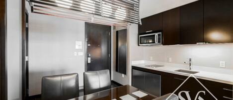 Una cocina moderna con armarios oscuros, un refrigerador de acero inoxidable y un microondas. Hay un comedor con una mesa de vidrio y sillas de cuero.