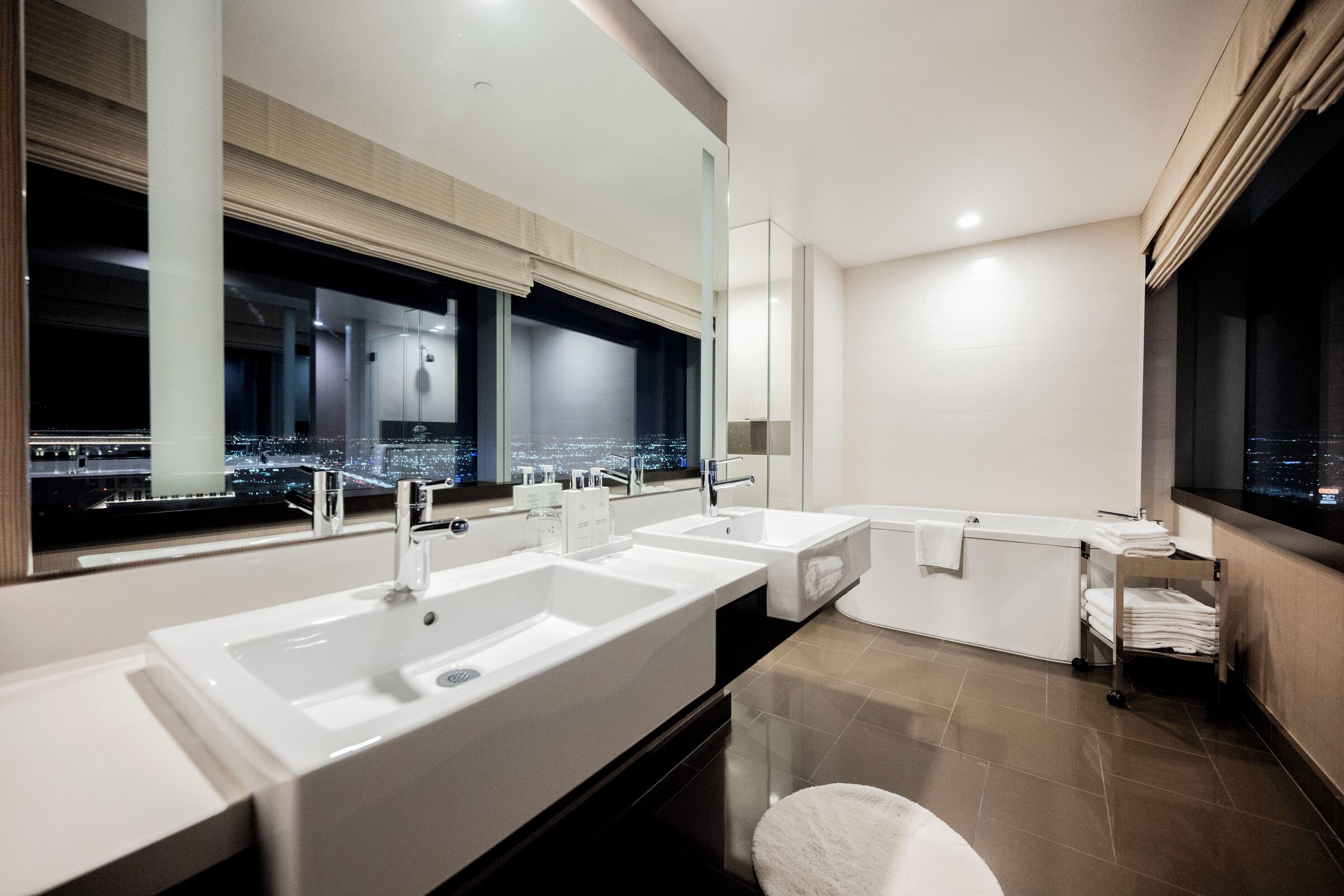 Un baño moderno con un tocador espacioso, dos lavamanos, una bañera y una ventana con vista a la ciudad.