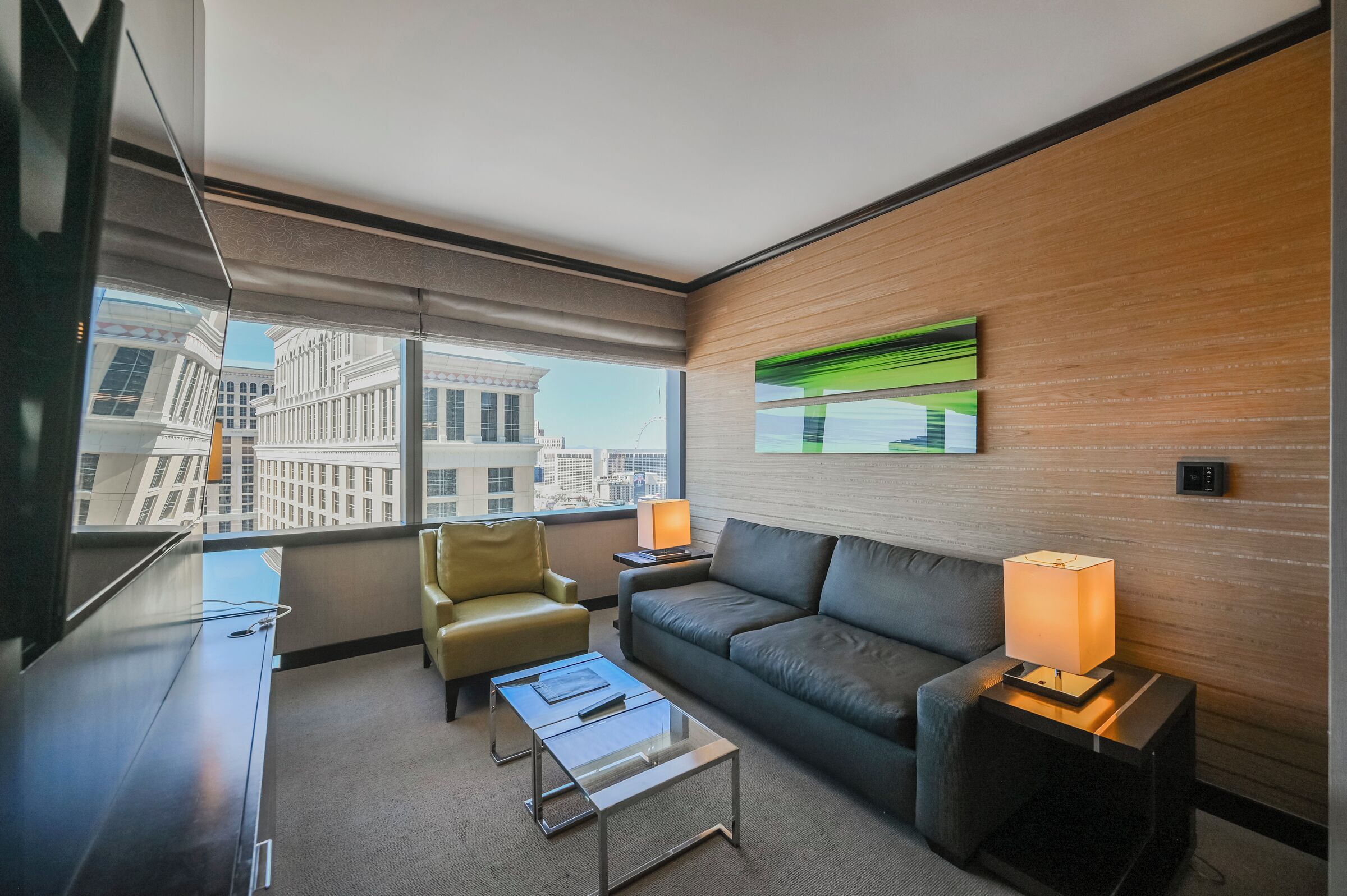 A modern hotel room with a large window, a leather sofa, a small armchair, a glass coffee table, and two bedside lamps.
