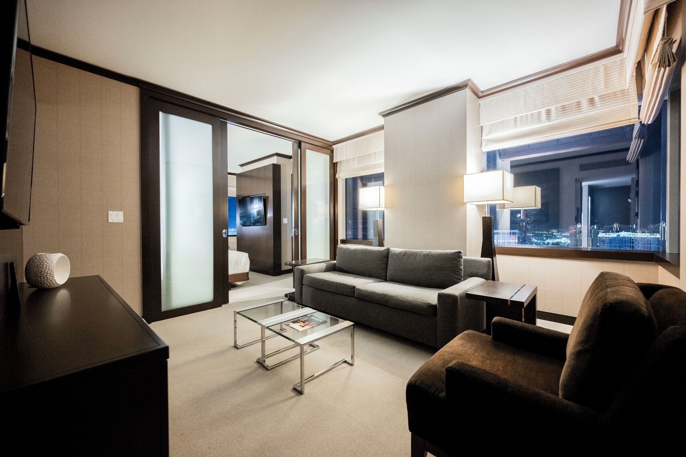 A modern hotel room with a sofa, armchair, and a glass coffee table. There is a city view through the window and a door leading to another room.