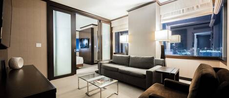 A modern hotel room with a sofa, armchair, and a glass coffee table. There is a city view through the window and a door leading to another room.