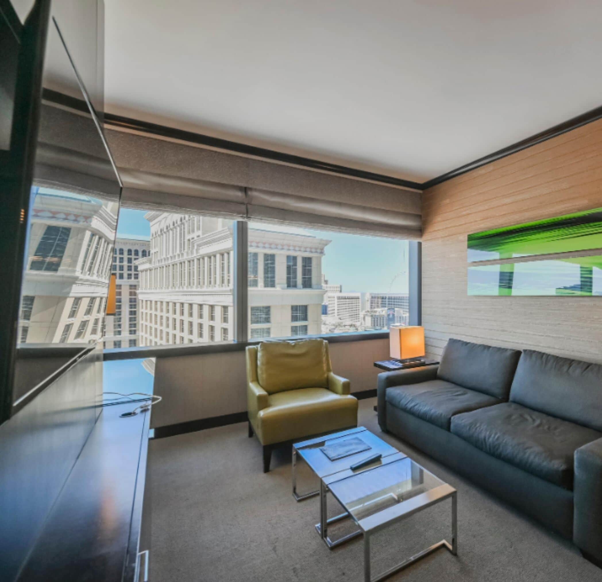 A modern hotel room with a large window offering a city view, a leather sofa, a leather armchair, and a glass coffee table.