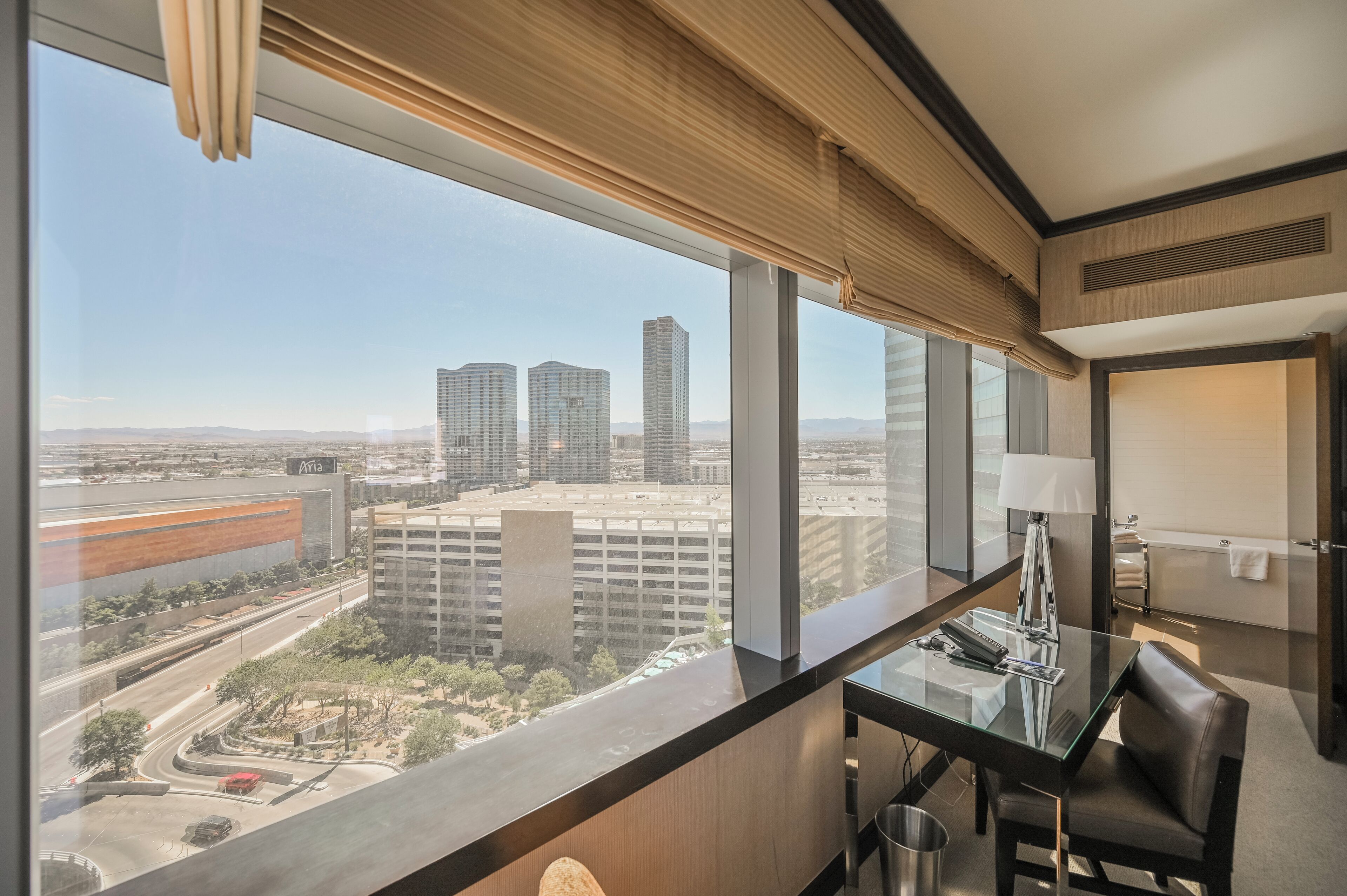 A hotel room with a large window offering a city view, a desk with a lamp, and a modern bathroom.