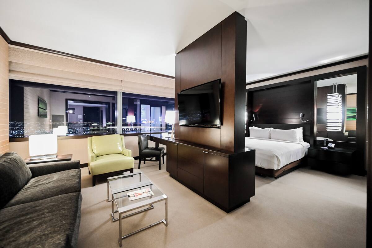Ett modernt hotellrum med en stor säng, en sittgrupp med soffa och fåtölj, en tv som är monterad på ett mörkt träskåp och utsikt över staden genom stora fönster.