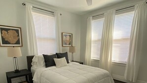 Ein Schlafzimmer mit einem Bett, einem Nachttisch, einer Stehlampe, einem Deckenventilator und großen Fenstern mit weißen Vorhängen.