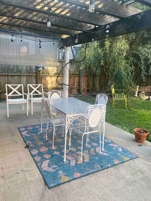 A covered patio area with a table and chairs, a blue patterned rug, and a wooden fence in the background.