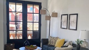 Ein Wohnzimmer mit einer dunkelblauen Tür, einem Deckenventilator, einem blauen Sofa mit gelben Kissen, zwei Sesseln, einem gemusterten Teppich und einem Beistelltisch mit Lampe und Pflanze.