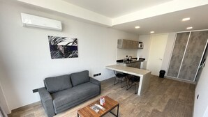 Ein moderner Wohnraum mit grauem Sofa, einem Holztisch und einer Küchenzeile mit Mikrowelle und Wasserkocher.