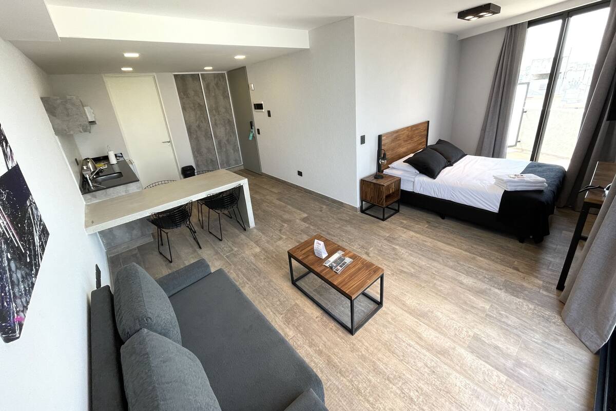 A modern hotel room with a bed, a sofa, a dining table, and a small kitchen area.