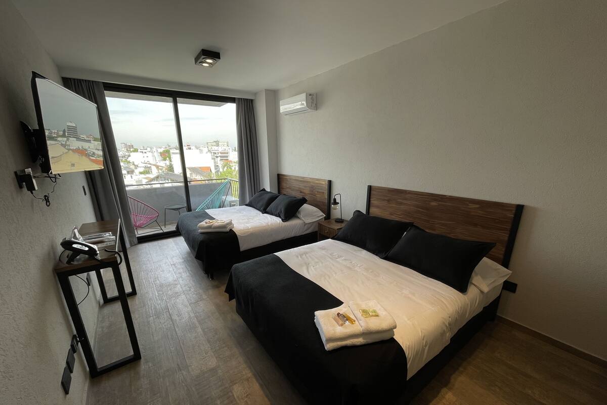 A hotel room with two beds, a TV, a desk, and a balcony with a city view.