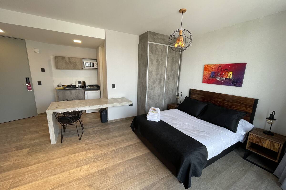 A modern hotel room with a bed, bedside table, and a kitchenette area.