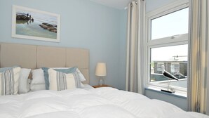 A neatly made bed with white bedding and striped pillows, a framed painting of a beach scene on the wall, and a window with a view of a building and a bird outside. - Slipway 16 (Wadebridge)