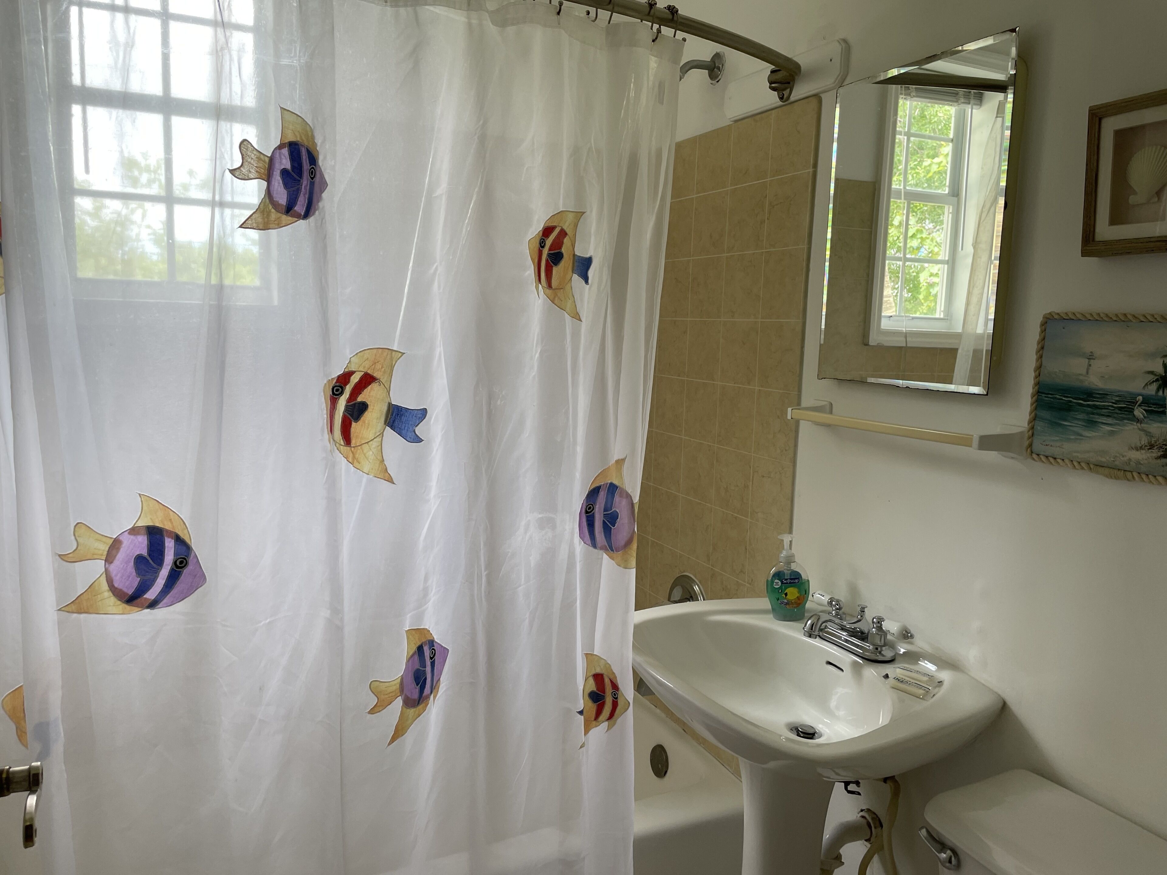 A bathroom with a shower curtain featuring colorful fish designs, a sink, a mirror, and a window with a view of greenery.