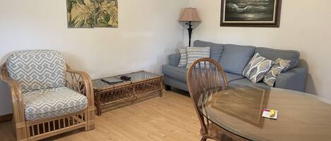 Una sala de estar con un sofá, dos sillones, una mesa de centro de vidrio, una mesa de comedor con sillas, una lámpara y dos obras de arte en la pared.