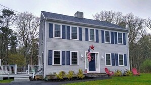 A two-story house with a front porch, a flag, and red Adirondack chairs. - Escape to the Cape in this brand new, family-friendly, centrally located home. (South Dennis)