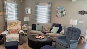 A living room with a sofa, armchairs, a coffee table, and a rug. There are two windows with curtains, a wall clock, and a small table with a lamp. - Escape to the Cape in this brand new, family-friendly, centrally located home. (South Dennis)