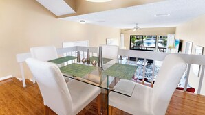 Comedor con mesa de vidrio, sillas blancas y vista a una piscina y un espacio exterior.