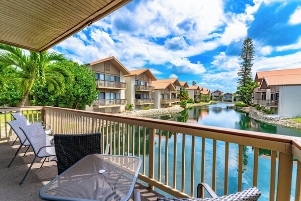 A balcony with wicker furniture overlooking a pool and a row of buildings.