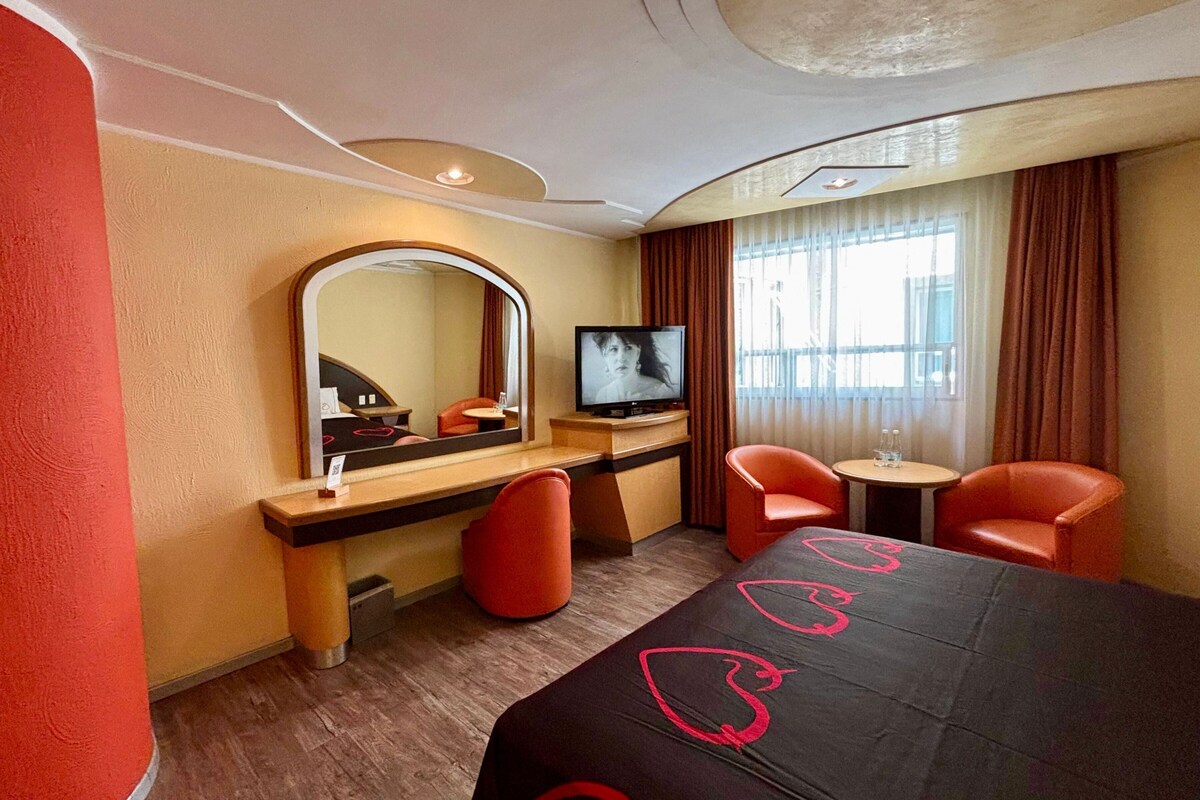 Une chambre d’hôtel équipée d’un lit, d’une télévision, d’un bureau, de deux chaises et d’un miroir.