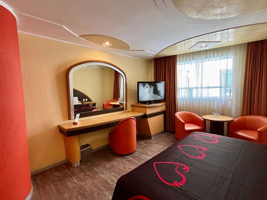 Una habitación de hotel con cama, televisión, escritorio, dos sillas y un espejo.