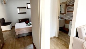Un dormitorio con cama, mesita de noche y un pequeño rincón de descanso. Se ve un acceso a un baño con inodoro y lavamanos.