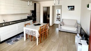 Una cocina moderna con armarios blancos, una mesa de comedor con sillas, un sofá y un televisor pequeño en la pared.