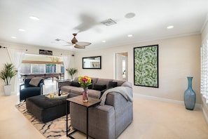 A modern living room with a grey sofa, a black armchair, and a coffee table. There is a ceiling fan, a large mirror, and a framed tropical plant on the wall.