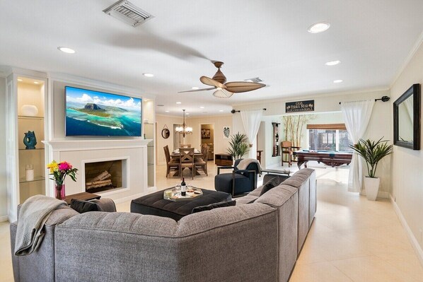 A spacious living room with a fireplace, a large flat-screen TV, a ceiling fan, and a dining area with a view of the city.