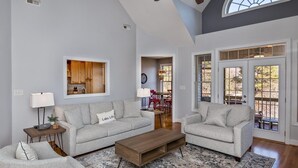 A spacious living room with two grey sofas, a wooden coffee table, and a patterned rug. A kitchen area is visible through an open door, and a dining area with red chairs is in the background. - Luxury in Rumbling Bald Resort ~ WiFi ~ Community Beach, Pool, Golf and more (Lake Lure)