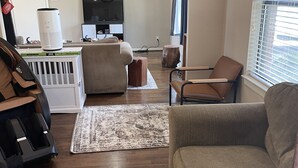 Una sala de estar con un sofá, una silla, una mesa de centro y un sillón de masaje. Hay un televisor en la pared y una ventana con persianas.