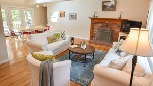 A living room with a fireplace, a sofa set, a coffee table, and a dining area with chairs. - Pond Front Home with Private Dock! (Brewster)