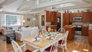 A spacious living area with a dining table set for six, a kitchen with wooden cabinets, and a large window providing natural light. - Pond Front Home with Private Dock! (Brewster)
