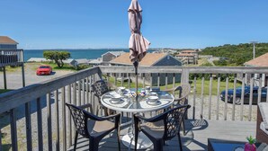 A deck with a table and chairs, overlooking a coastal area with buildings and a red car in the distance. - Views of Bay Access to Private Beach! (North Truro)
