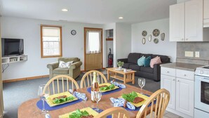 A dining area with a wooden table set for four, a kitchen with white cabinets, and a living room with a dark sofa and decorative plates on the wall. - Views of Bay Access to Private Beach! (North Truro)