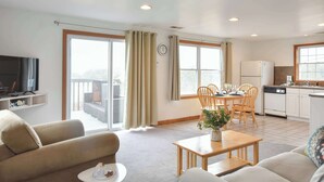 A modern living room with a sofa, coffee table, and dining area with chairs. A television is mounted on the wall, and there is a kitchen area with a refrigerator and dishwasher. - Views of Bay Access to Private Beach! (Truro)