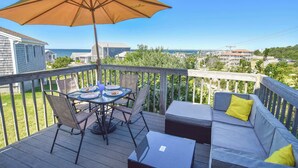 A wooden deck with a table and chairs, a sofa with cushions, and an umbrella. - Views of Bay Access to Private Beach! (Truro)