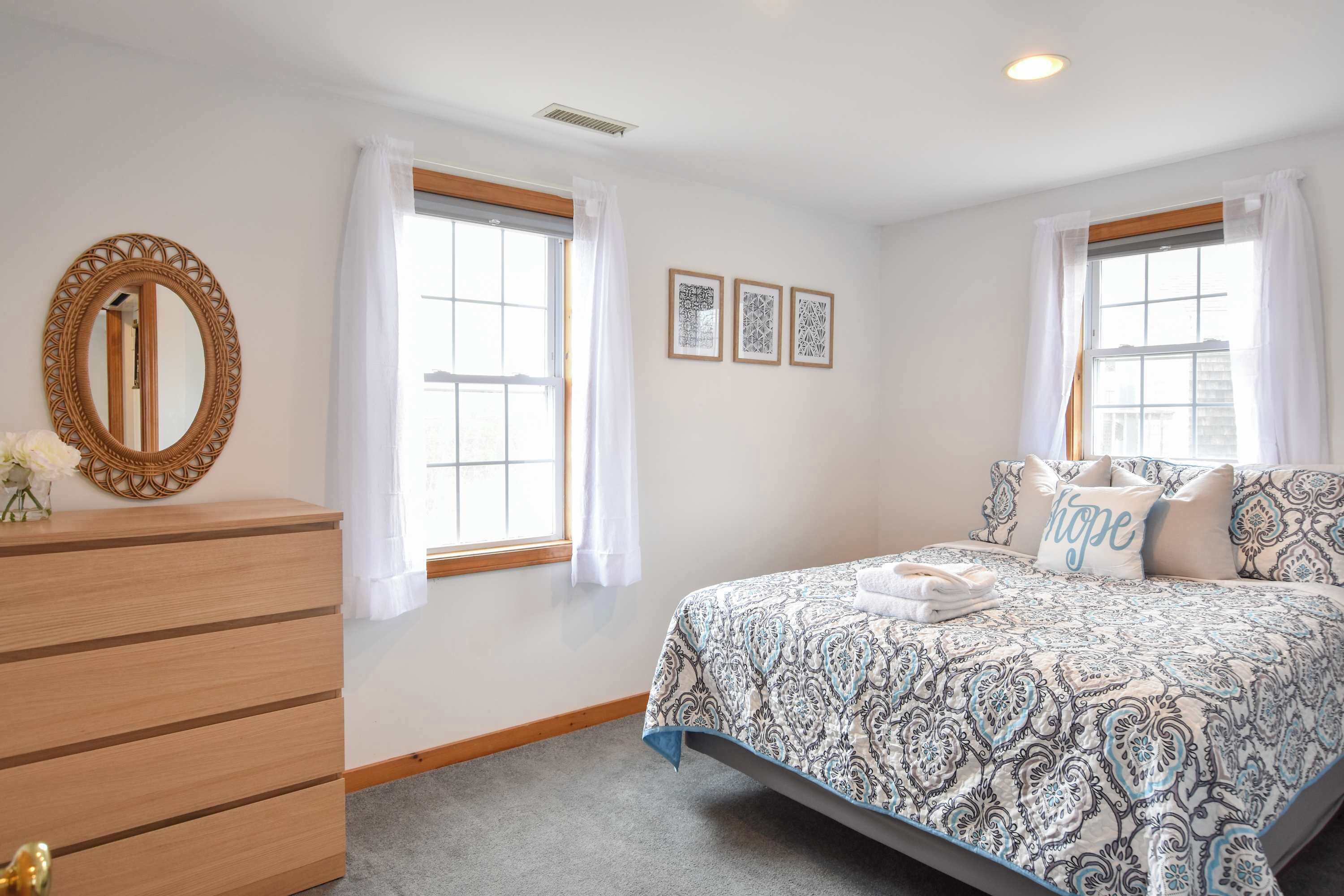 A bedroom with a bed, a dresser, a mirror, and two windows with curtains.